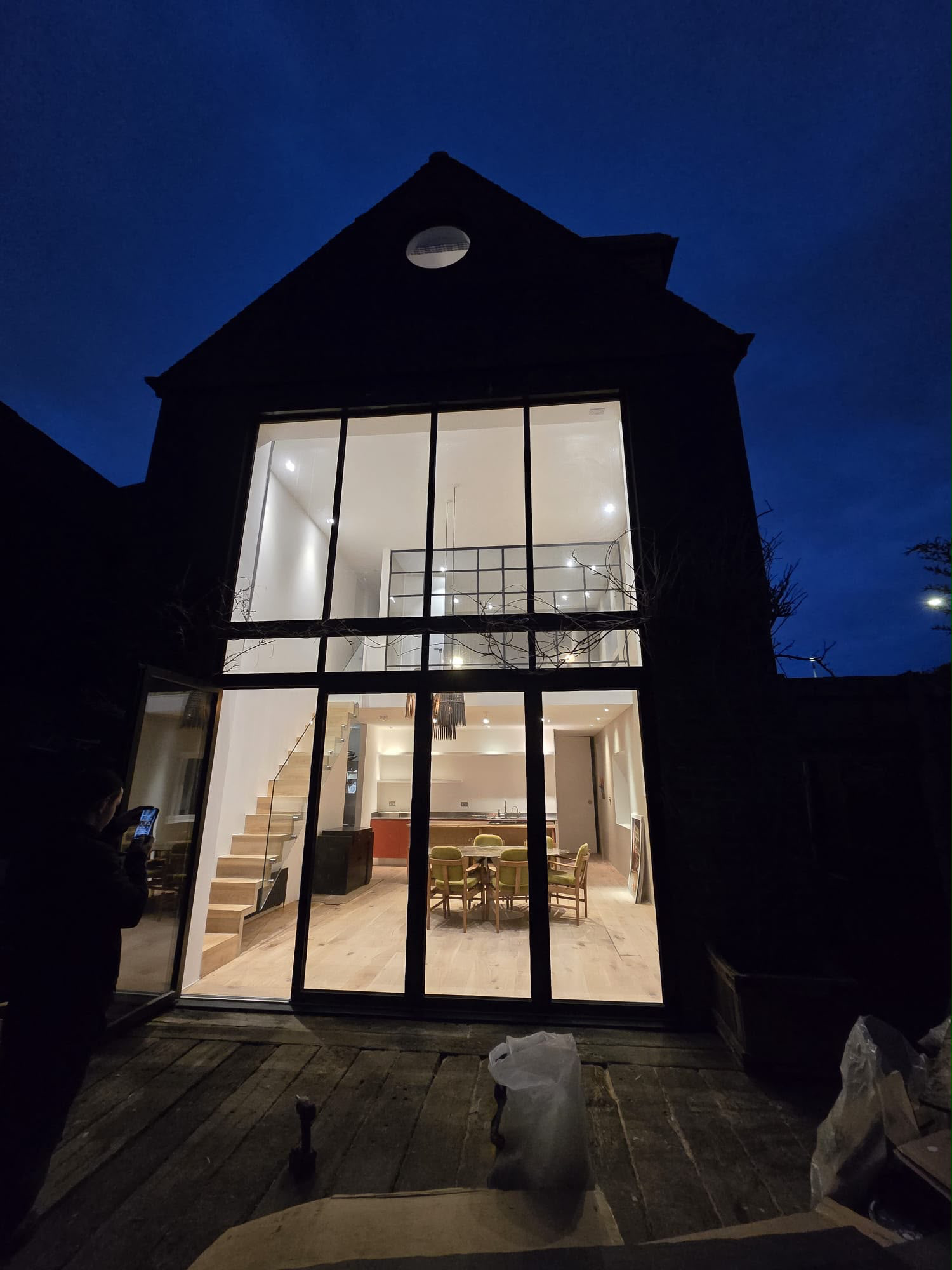 Tall glazed rear elevation at dusk revealing a double-height dining space, mezzanine level and circular roof opening