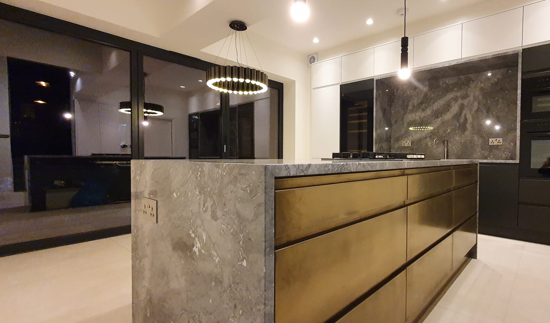 Bespoke marble kitchen island with brushed brass cabinetry, ring pendant light and full-height glazing onto the garden